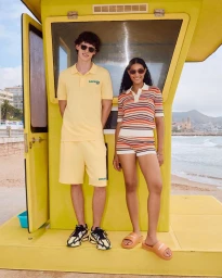 Two models wearing yellow Lacoste apparel and sunglasses posing in front of a bright yellow lifeguard tower on a sandy beach.