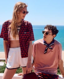 Two models wearing Lacoste summer apparel outdoors by the ocean, with a bright blue sea and clear sky backdrop.