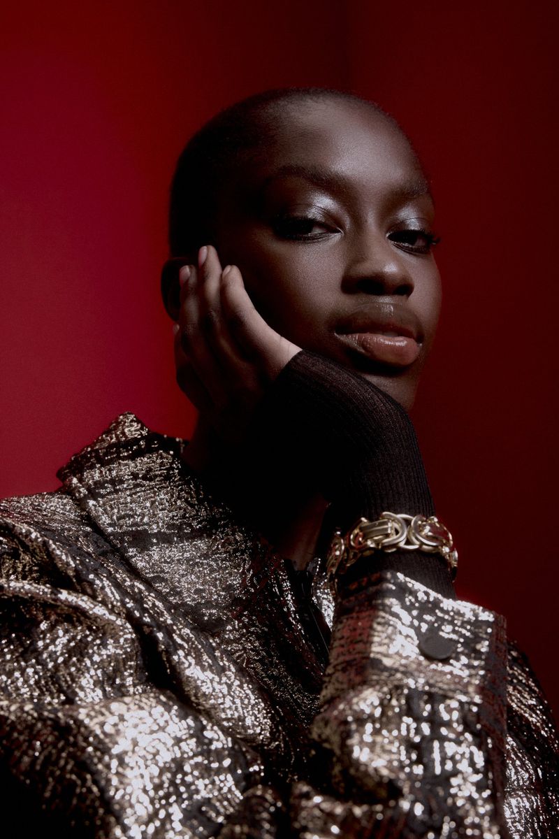 Portrait of a young adult African woman with bare head. She is wearing a gold sequined jacket, leaning on her hand, set against a red background.