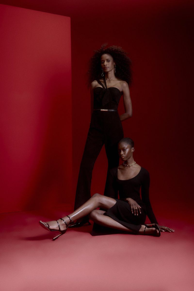 Two Black fashion models strike poses in a red photo studio. One woman stands tall; the other sits on the floor in a long sleeve dress.