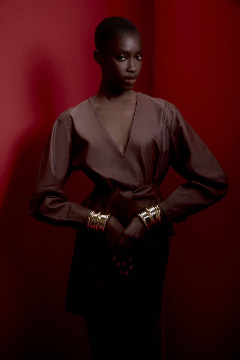 Portrait of an African American woman in a dark wrap dress with gold bracelets, posing against a deep red studio backdrop.