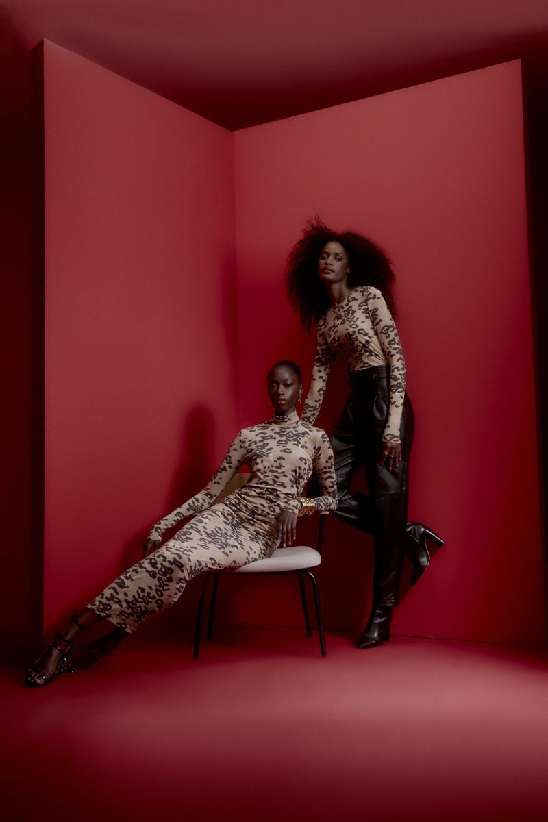 Two Black women pose in matching beige and black animal print tops in a red-walled corner. One is seated; the other stands in high-heeled boots.
