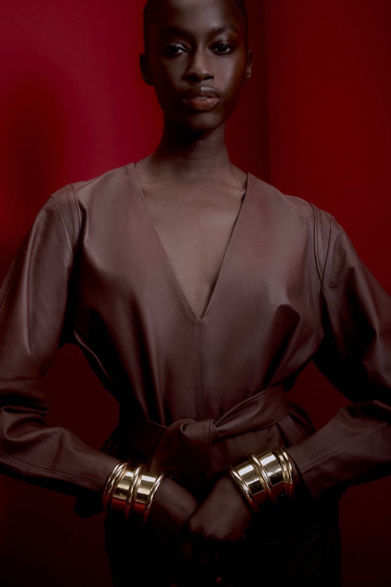 Headshot of an African American woman in a brown dress with long sleeves, a v-neck, and a matching belt, posing against a red background.