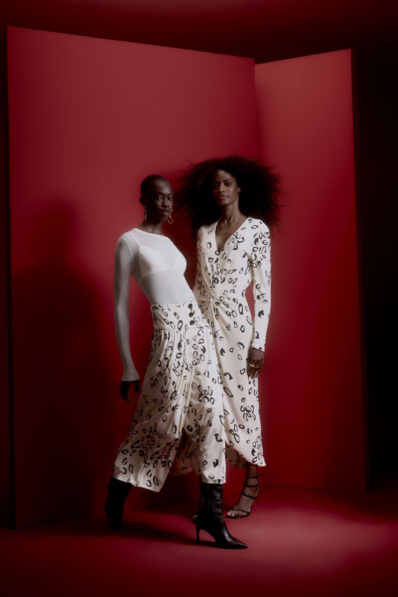 Two adult women pose in a red studio wearing long-sleeved, patterned dresses and black boots. One woman wears a sheer top.