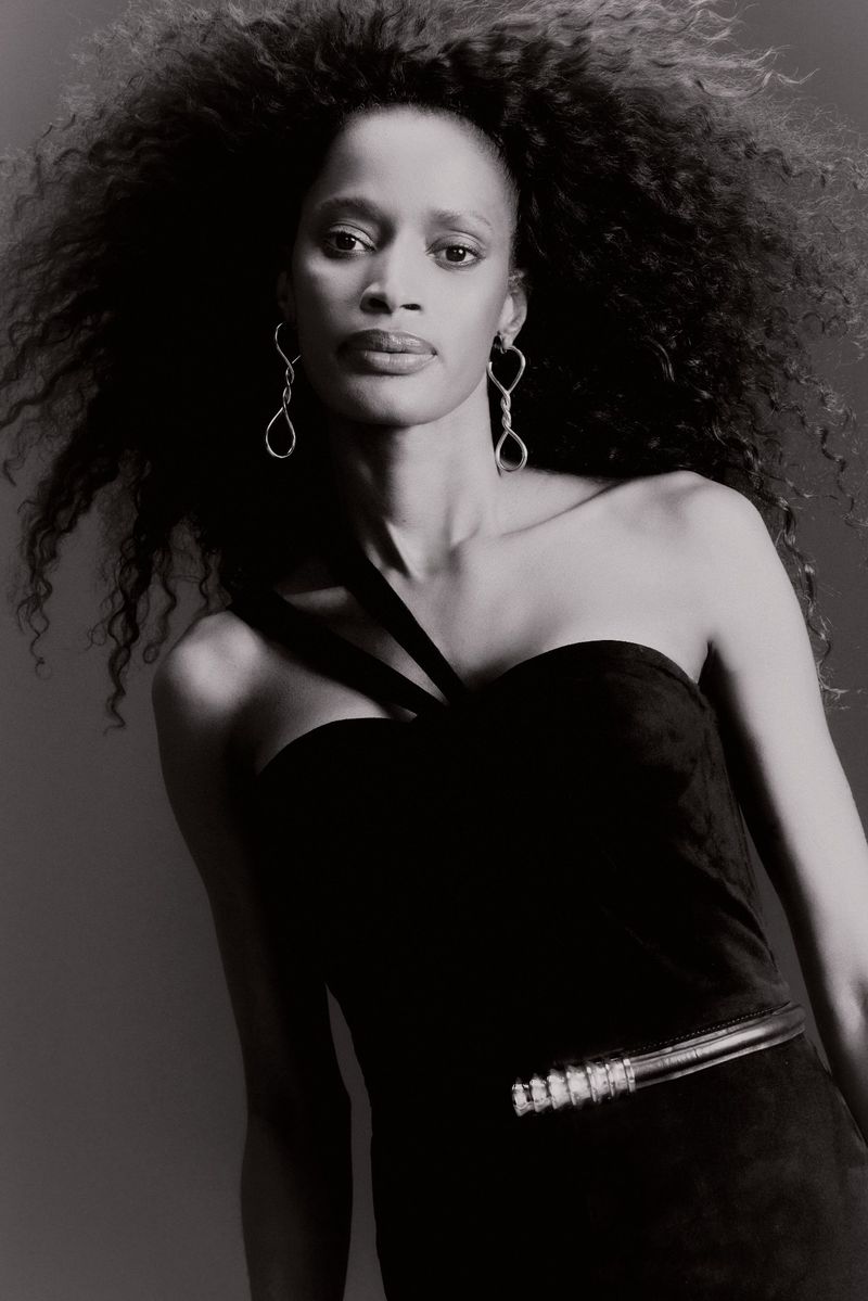 Black and white portrait of an adult female with curly hair wearing a black one-shoulder dress and statement earrings against a grey background.