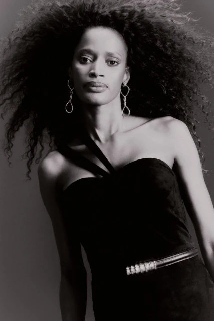 Black and white portrait of an adult female with curly hair wearing a black one-shoulder dress and statement earrings against a grey background.