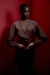 Model with a shaved head wearing a belted brown top and gold cuffs against a deep red background for Animale