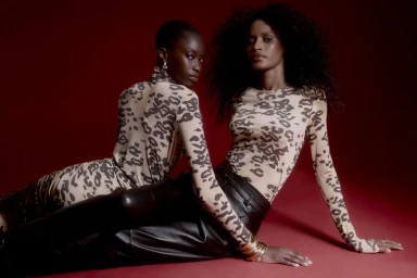 Two black women models wearing leopard print tops and black trousers posing together on a dark red studio floor