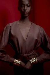 Model wearing a brown belted top with thick gold cuffs against a deep red background for the Animale campaign