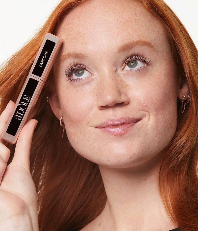 Red-haired woman holds Lancôme Idôle mascara, showcasing beauty product and makeup routine in a clean, close-up beauty shot.
