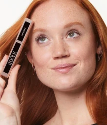 Red-haired model holding a Lancôme Idôle mascara tube near her face against a neutral background