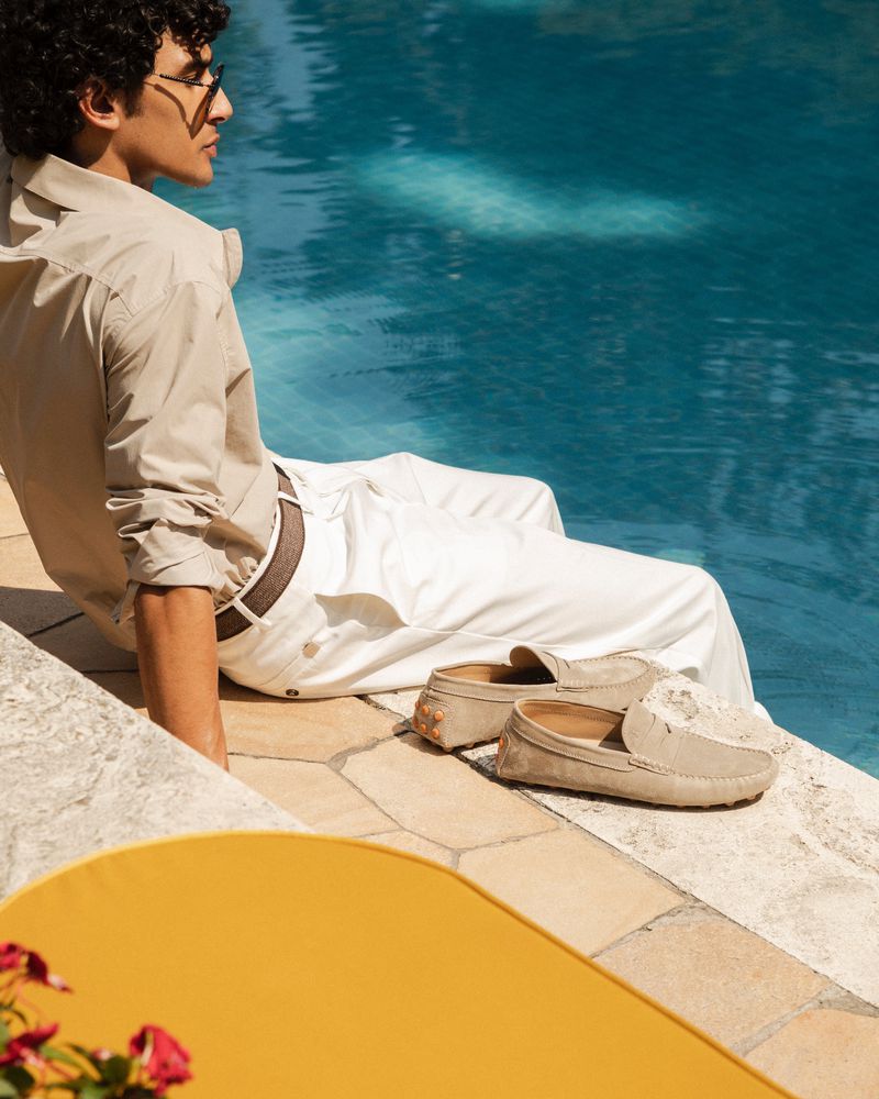 A man wearing sunglasses and light-colored clothes sits beside a pool with beige suede loafers, offering a relaxed and stylish scene.