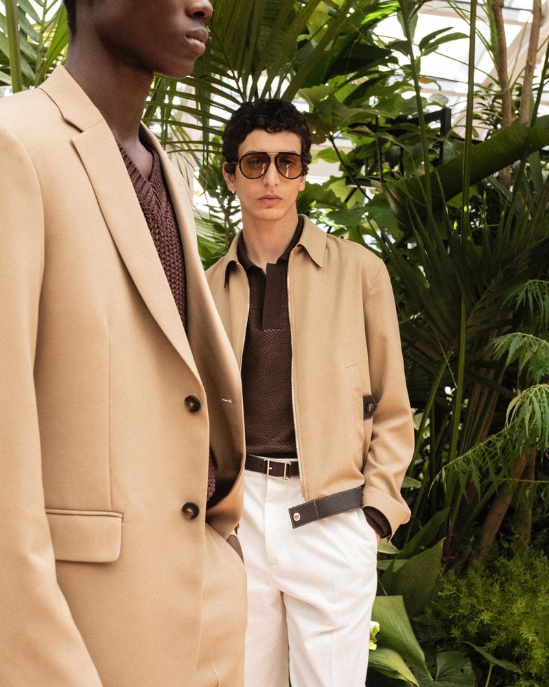 Two adult men pose in a stylish outdoor fashion shoot, wearing a tan blazer and a jacket paired with white pants against a leafy backdrop.