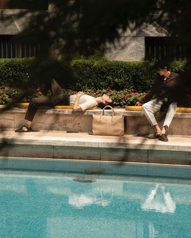 Two people, one lying down and the other sitting, are relaxing on a bench next to a swimming pool on a sunny day.