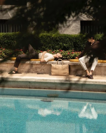 Two people, one lying down and the other sitting, are relaxing on a bench next to a swimming pool on a sunny day.