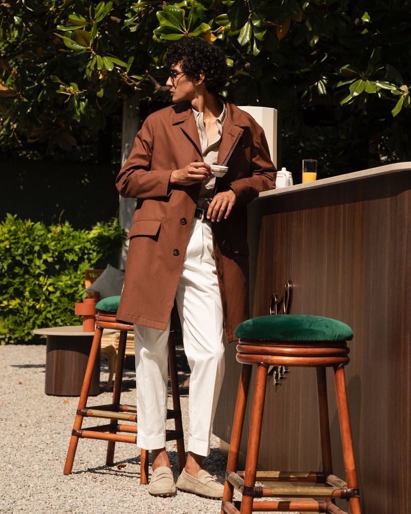 Man with sunglasses stands in a brown coat next to tall bar stools, holding a cup. Outdoor patio setting with greenery.