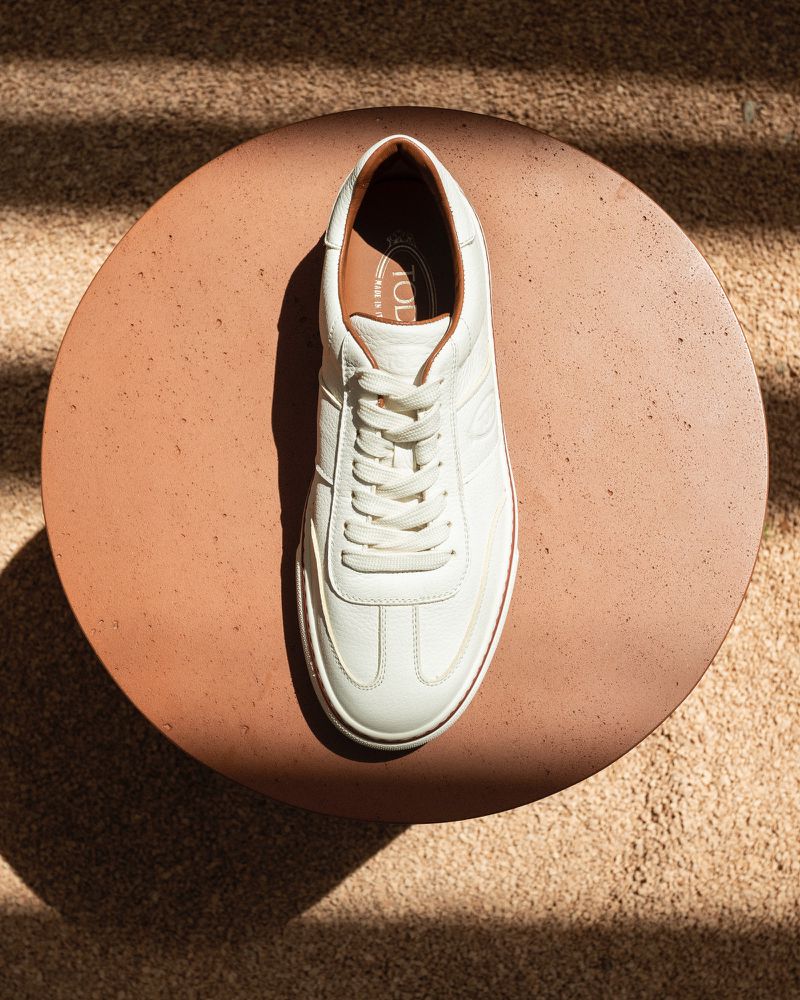 Elevated view of a white leather Tod's sneaker on a round table with a carpeted surface, captured in soft, natural lighting.