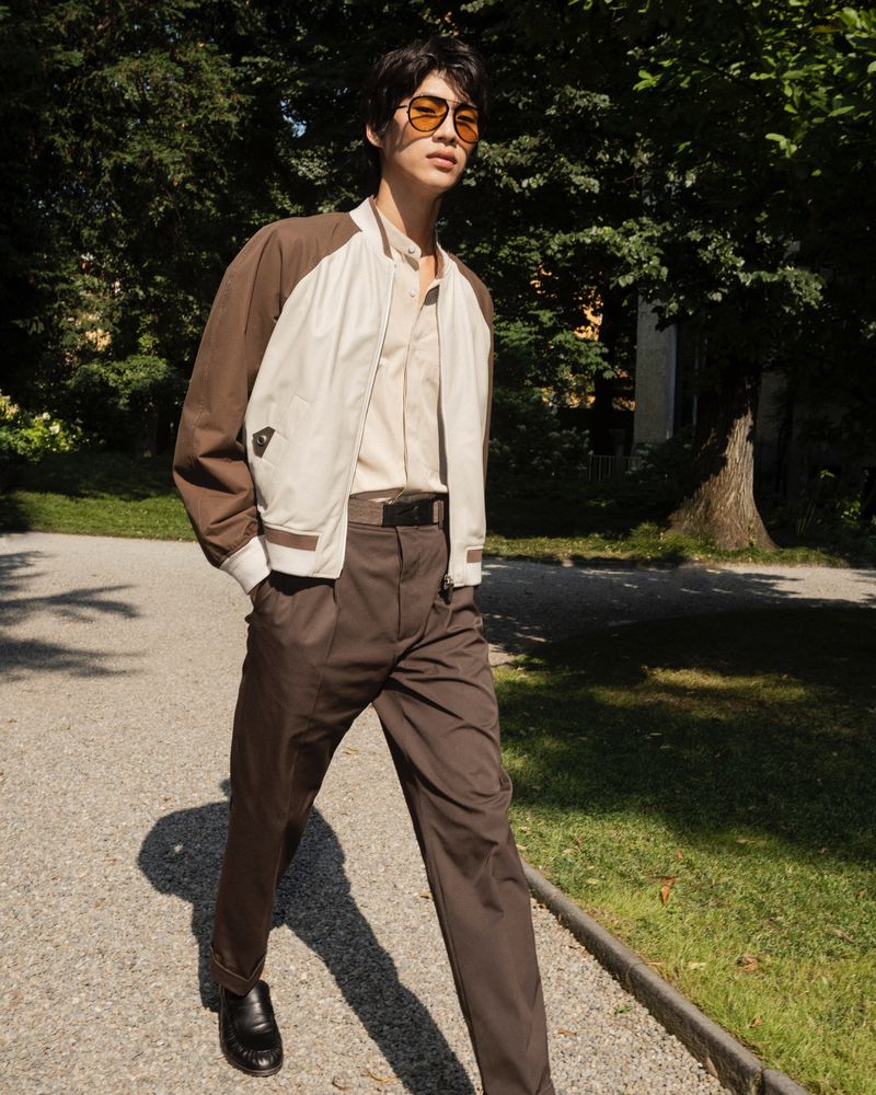 Adult male in bomber jacket and brown trousers walking in park setting with trees and shadows, wearing sunglasses and dress shoes.
