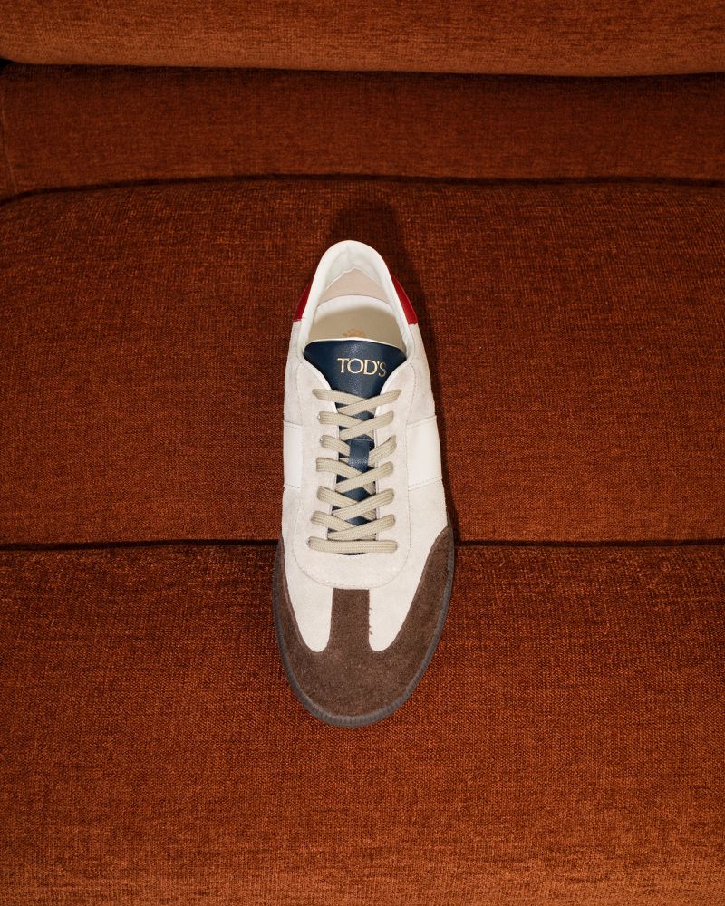 Close-up of a cream-colored Tod's sneaker with brown toe and gray laces placed on a ribbed brown textile surface.