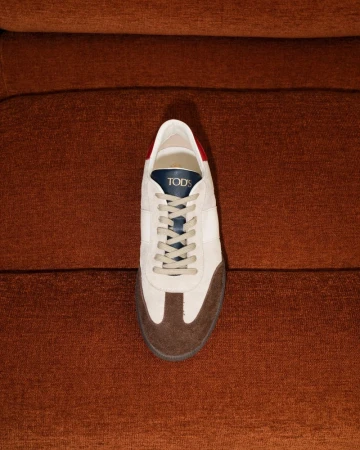 Close-up of a cream-colored Tod's sneaker with brown toe and gray laces placed on a ribbed brown textile surface.