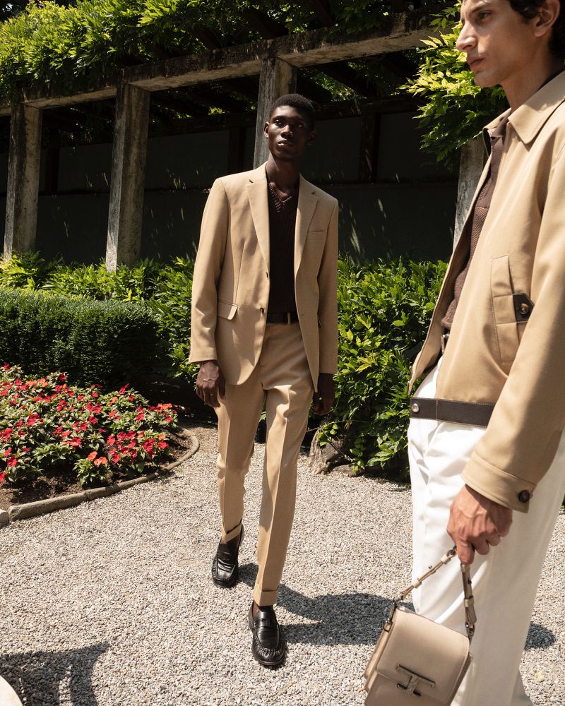 Two adult men in tan suits stand outdoors on a gravel path among green foliage, creating a sophisticated and classic fashion scene.