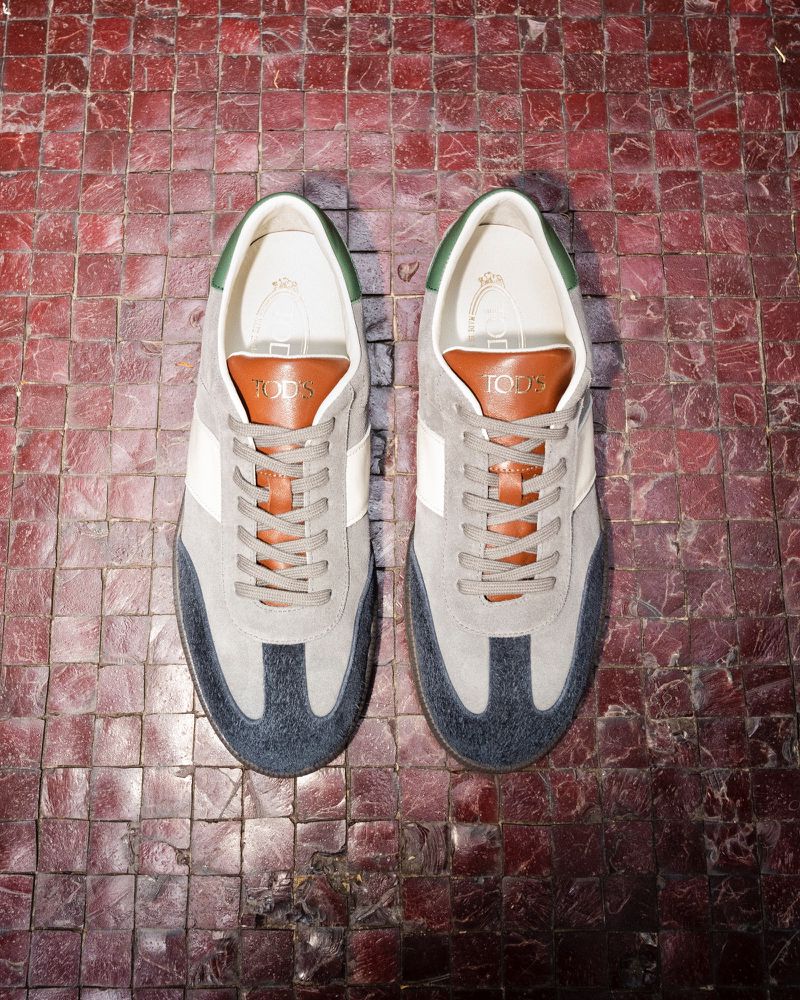 Overhead product shot of a pair of Tod's suede sneakers, gray with white and navy details, and tan leather tongue patch, on a red tile background.