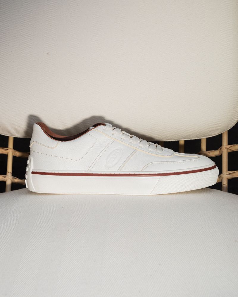 A white leather sneaker with brown accents sits on a light-colored cushioned chair, showcasing a classic and clean athletic aesthetic.