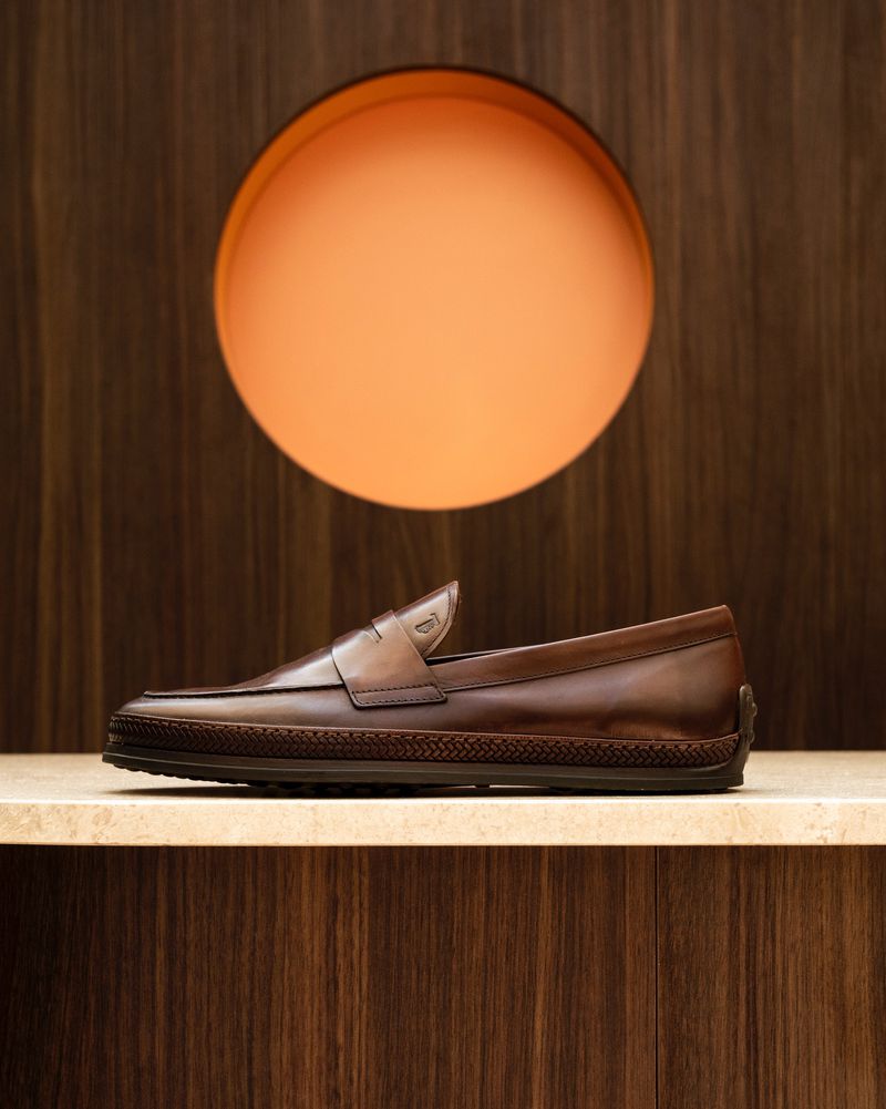 Brown leather loafer shoe displayed on a marble shelf against a wood-paneled wall with a circular orange light fixture.