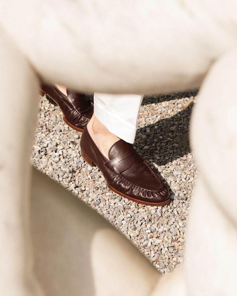 Leather penny loafers on a person standing on a gray pebble surface next to a textured white architectural element outside.