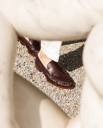 Leather penny loafers on a person standing on a gray pebble surface next to a textured white architectural element outside.