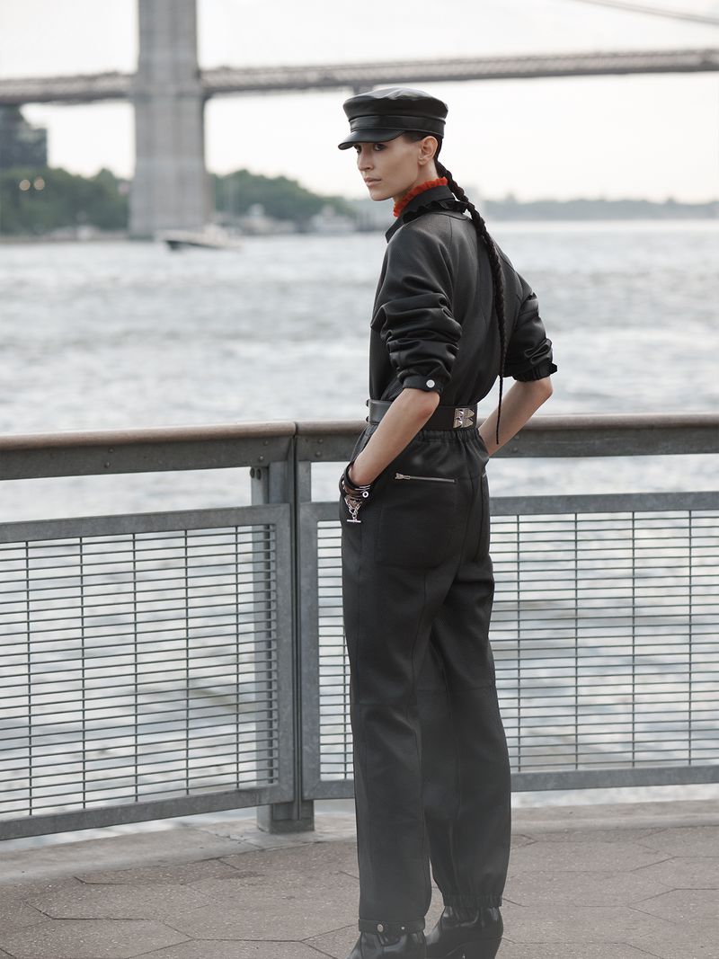 A person in a dark jumpsuit and cap poses in front of a waterfront, showcasing modern fashion with an urban backdrop and natural light.