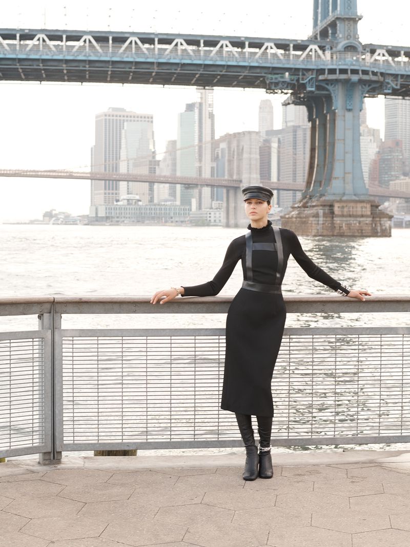 Woman in black dress, boots, and hat poses near waterfront railing with bridge and city skyline in background on a bright day.