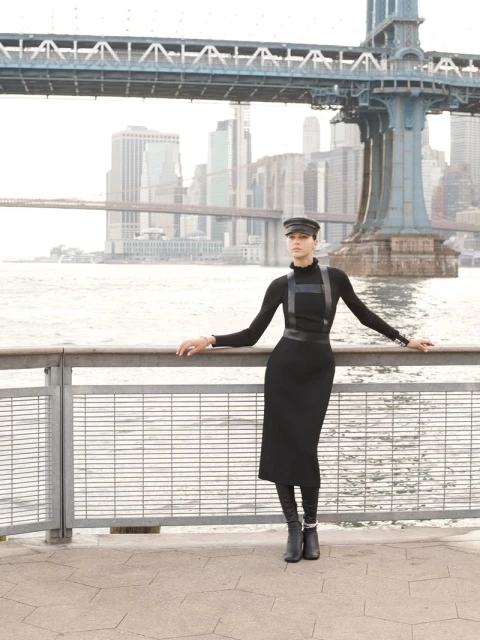 Woman in black dress, boots, and hat poses near waterfront railing with bridge and city skyline in background on a bright day.