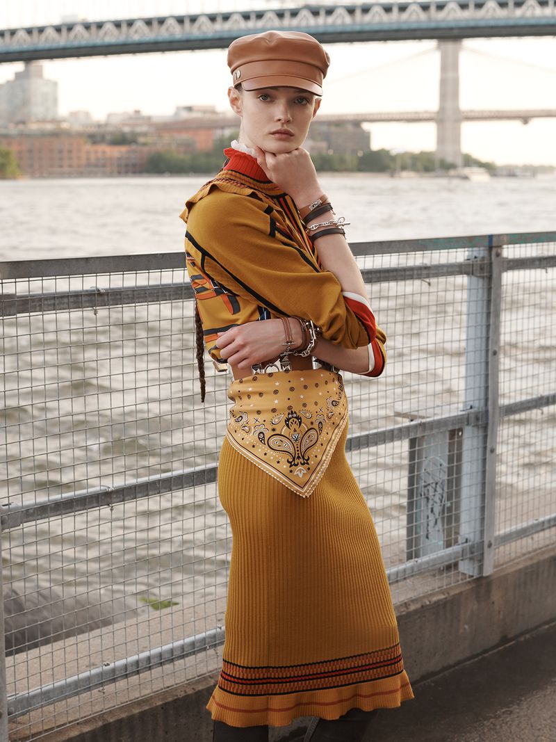 Adult woman poses in a mustard midi-dress, leather cap, and bandana belt, with a metal guardrail and a city bridge in background.