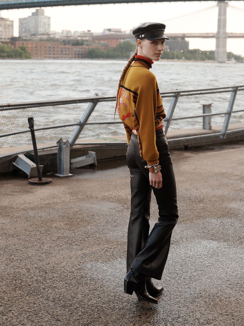 Woman in black leather pants and cap poses near river with bridge in background, showcasing fashion and urban style.