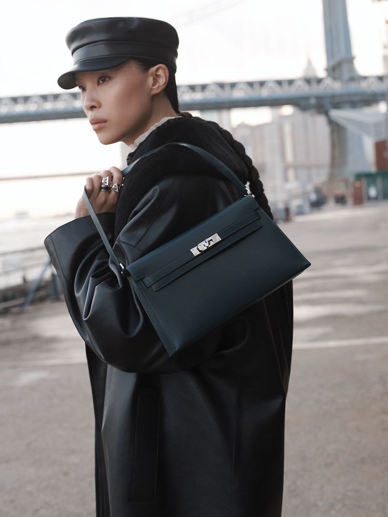 Asian woman in a hat and black coat carrying a structured black leather handbag on a city street with a bridge and buildings.