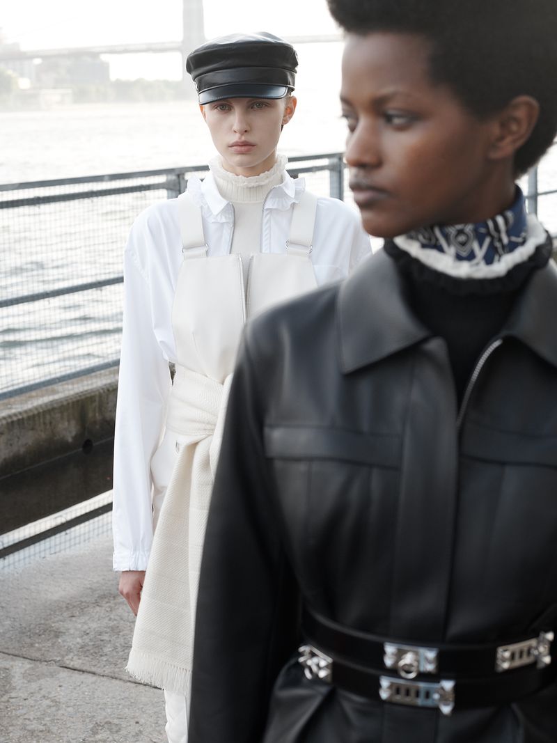 Two fashion models pose outdoors on a concrete surface. One model wears a black leather jacket and the other a white top and cap.