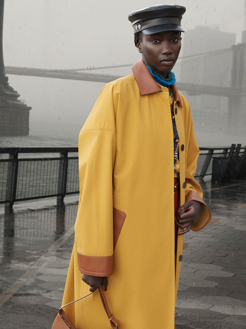 A woman in a yellow coat stands outdoors against a backdrop of a bridge on a grey day, carrying a tan purse. Fashion editorial.