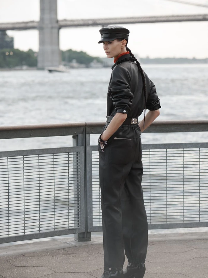 Model in black leather jumpsuit and cap standing by waterfront railing with suspension bridge in background