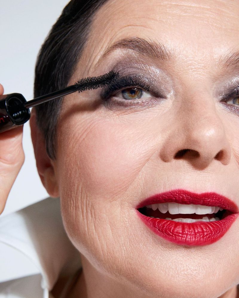 Close-up of an adult woman applying black mascara to her eyelashes while wearing red lipstick in a bright indoor setting.