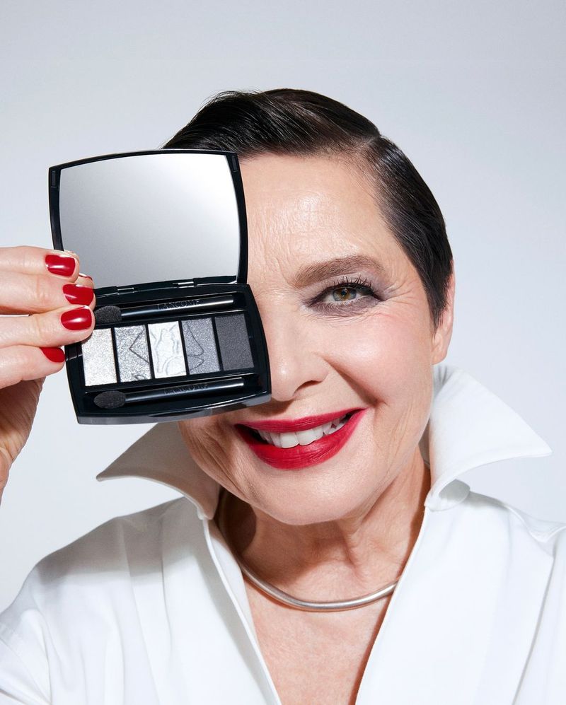 Mature woman smiles holding a gray eyeshadow palette over her eye in a studio shot, showcasing makeup and a clean, white collared shirt.