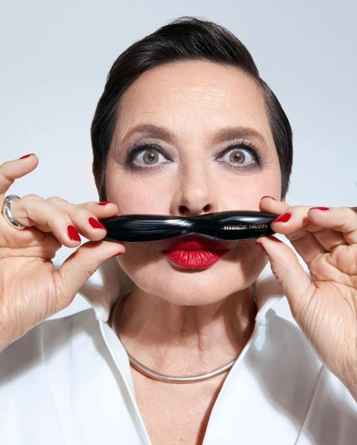 Woman holds black mascara tube as a mustache, smiling with red lipstick in an indoor photo studio against a white backdrop.