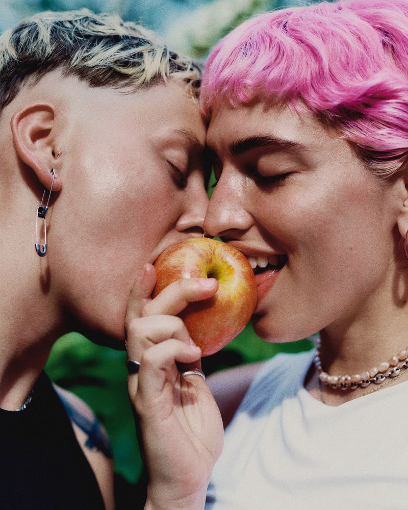 Close-up of two people sharing an apple, showcasing their facial features and hairstyles; one has pink hair and pearl necklace.