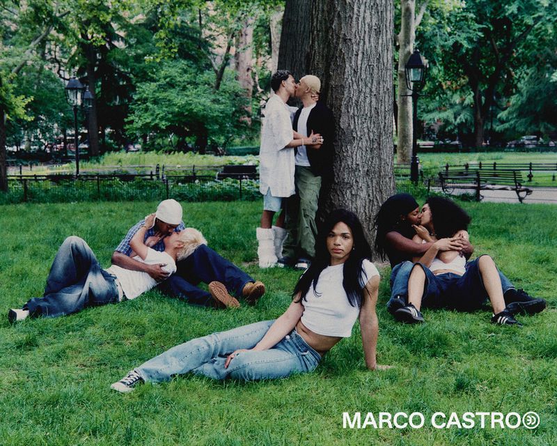 A diverse group of couples gather outdoors in a grassy park setting. Some are standing while others are sitting or lying down on the green grass.