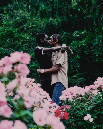 Two Black men embrace and kiss in a garden with pink flowers in the foreground and greenery in the background, captured in natural light.