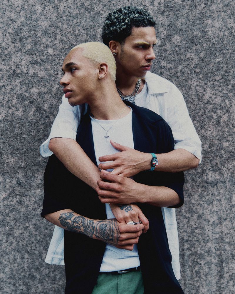 Two adult men pose in a medium shot with a neutral facial expression in front of a gray textured wall, wearing casual clothing.