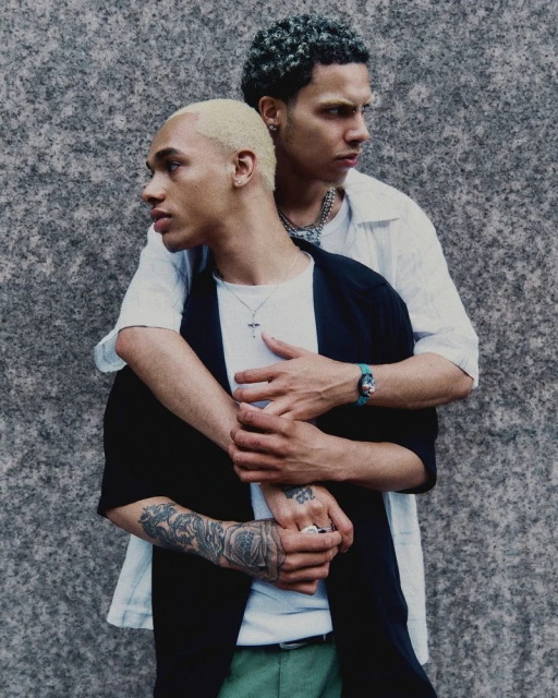 Two adult men pose in a medium shot with a neutral facial expression in front of a gray textured wall, wearing casual clothing.