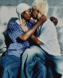 Two men embracing and kissing outdoors while seated against a stone wall for the MARCO CASTRO Pride Campaign.
