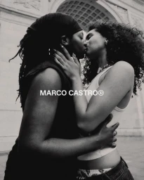 Black and white portrait of two women kissing against a stone architectural backdrop for the Marco Castro Pride Campaign.