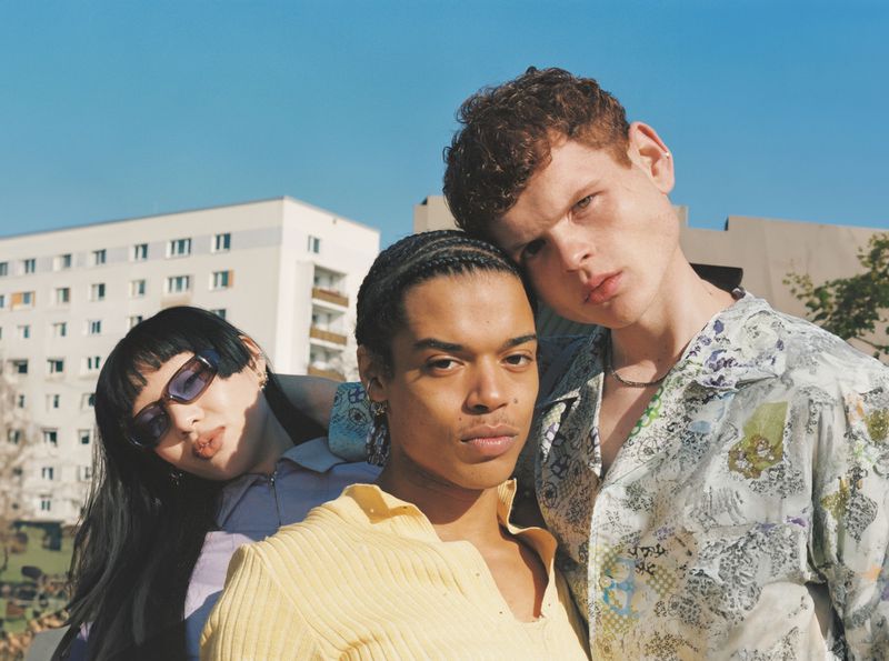 Three models posing closely together outdoors in front of a residential building under a clear blue sky for RIMOWA.